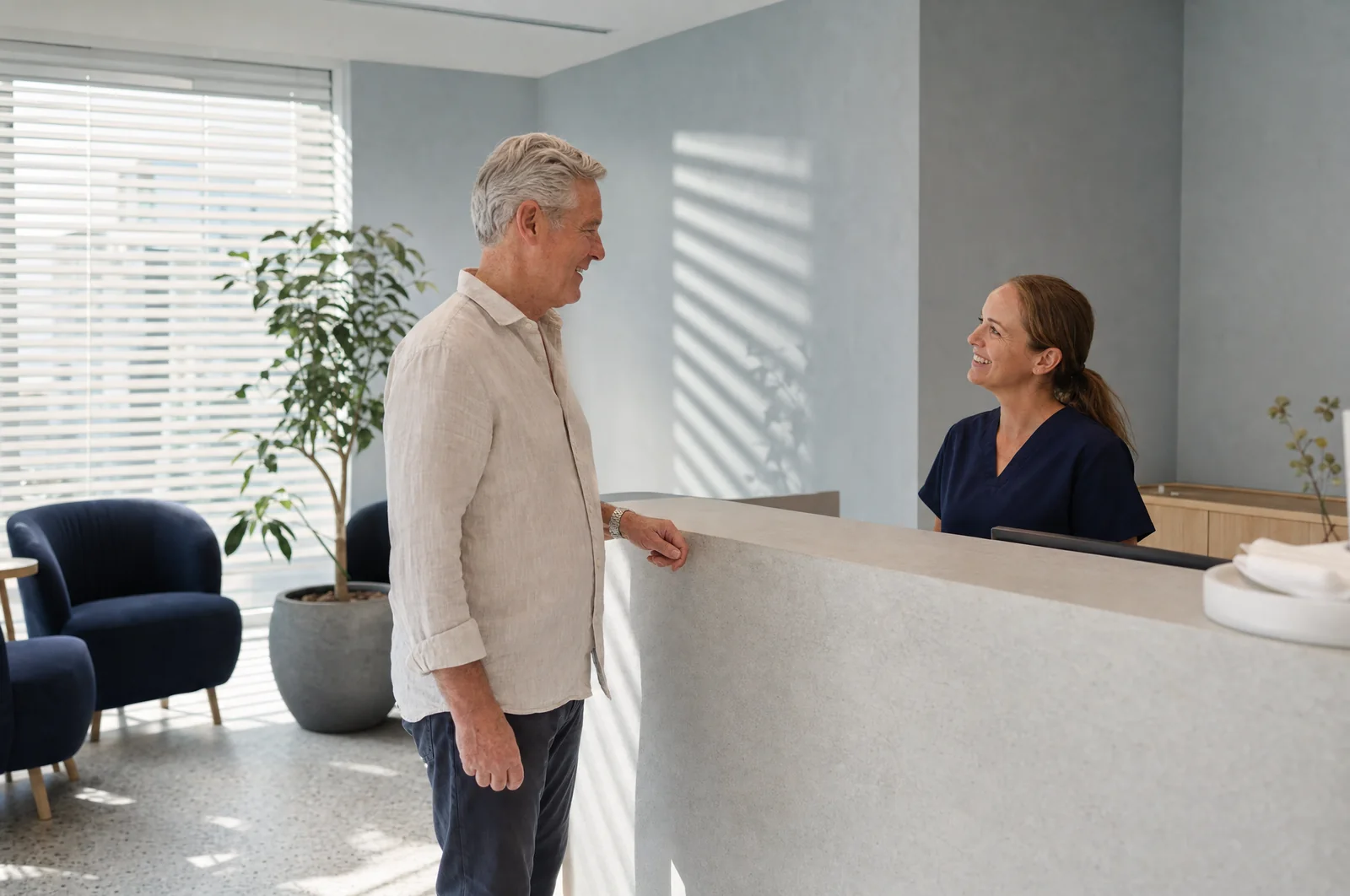 Illustrative scene: a man in a linen shirt talks with a smiling receptionist in navy scrubs across a pale stone front desk, armchairs and blinds behind him.