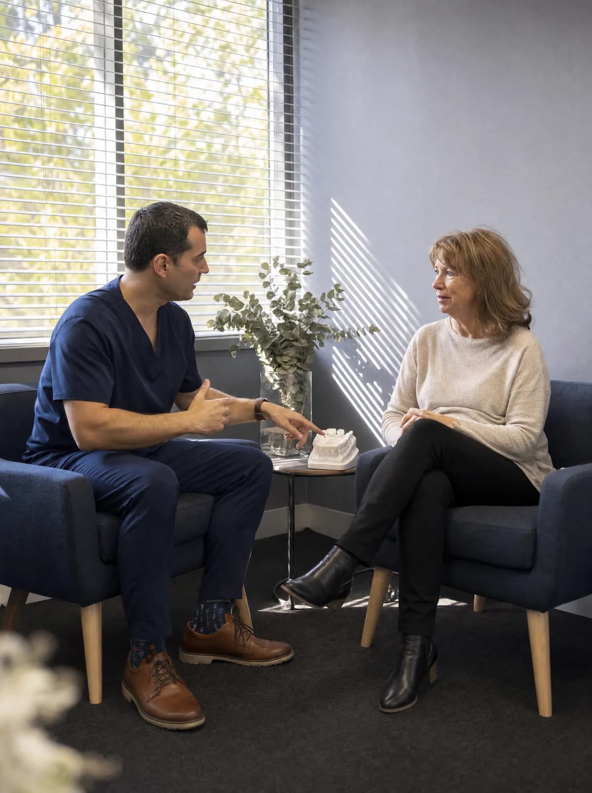 Illustrative scene: a dentist in navy scrubs and a woman sit in two armchairs by a window with blinds, talking over a white model of teeth.