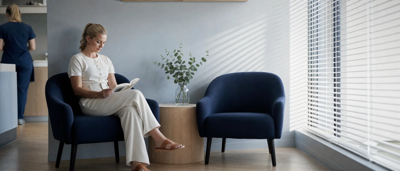 Illustrative scene: a woman reads in a quiet waiting lounge with navy armchairs, a eucalyptus sprig on an oak side table and long stripes of light from the blinds.