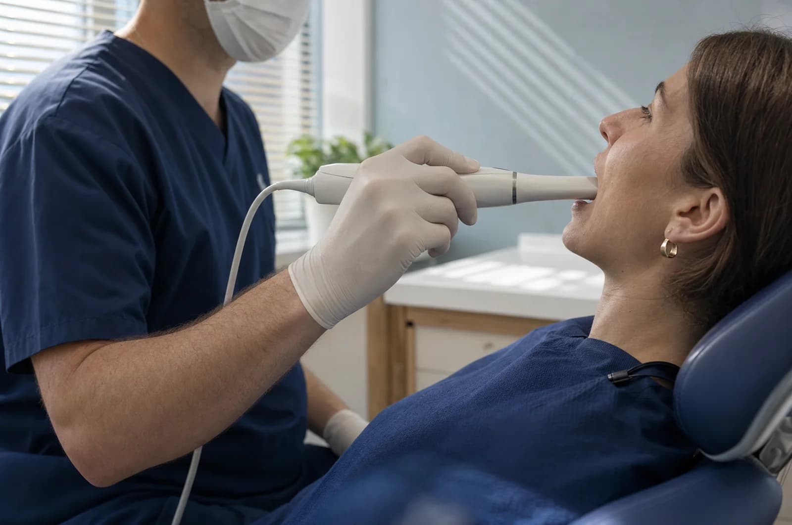 Illustrative close-up: a dentist in navy scrubs holds a slim intraoral scanner at the lips of a patient reclined in a navy dental chair.